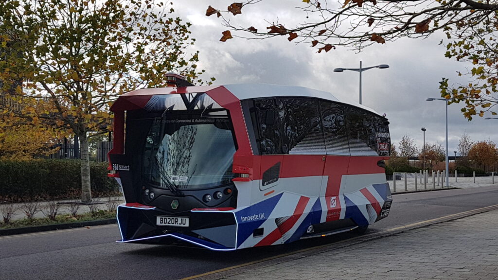 An autonomous bus, with a union jack flag on the side of it, on the road in Cambridge