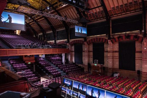 Photo shows the inside view of the Cambridge Corn Exchange - tiered seating in the venue and digital screens hanging from the ceiling with some computer screens in the foreground.