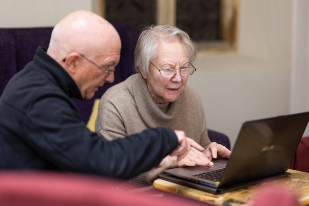 Two older people sitting at a table using a laptop, with one person pointing at the screen while the other types.