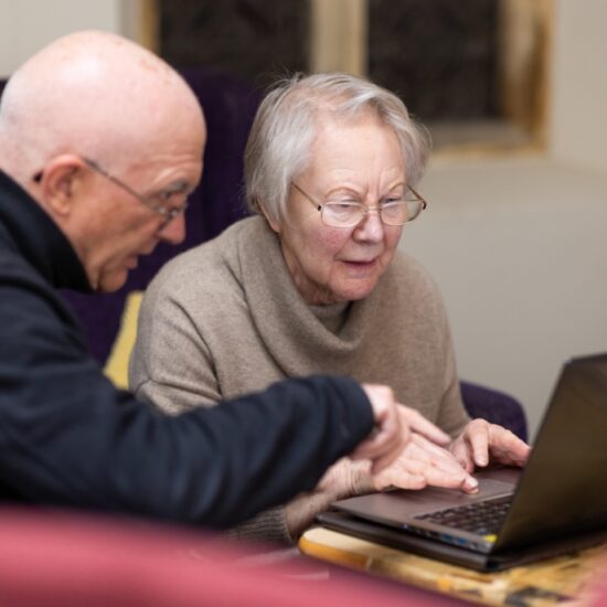 Two older people sitting at a table using a laptop, with one person pointing at the screen while the other types.