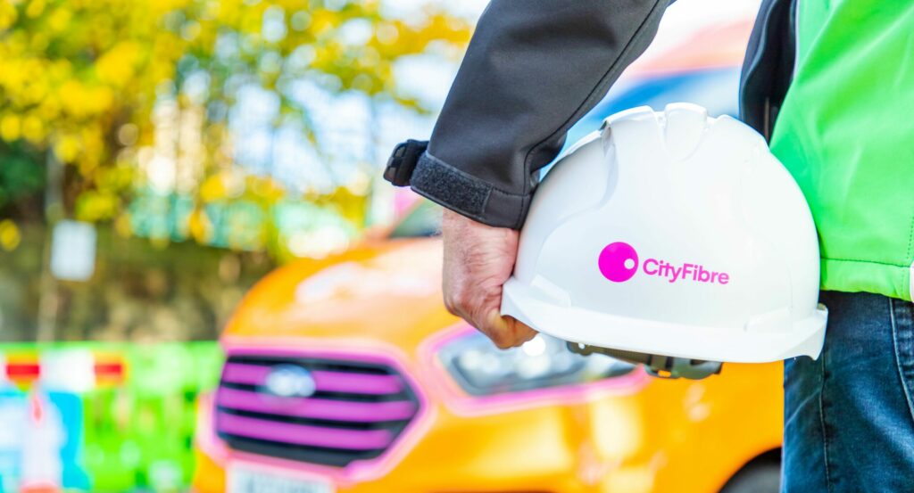 Photo of someone's arm holding a white, hard hat with the CityFibre logo on it.
