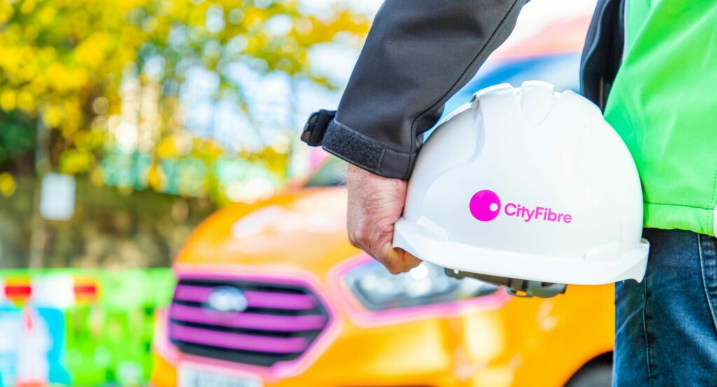 Photo of someone's arm holding a white, hard hat with the CityFibre logo on it.