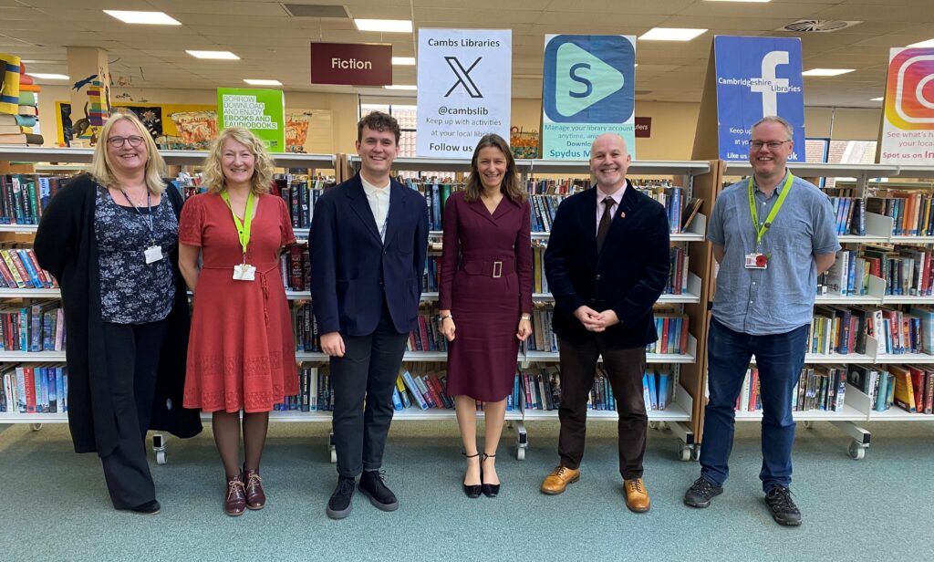 Lucy Frazer MP in Ely Library with representatives from Connecting Cambs, Cambs Libraries & Cambs Skills. Everyone is standing in a row, smiling by bookshelves.