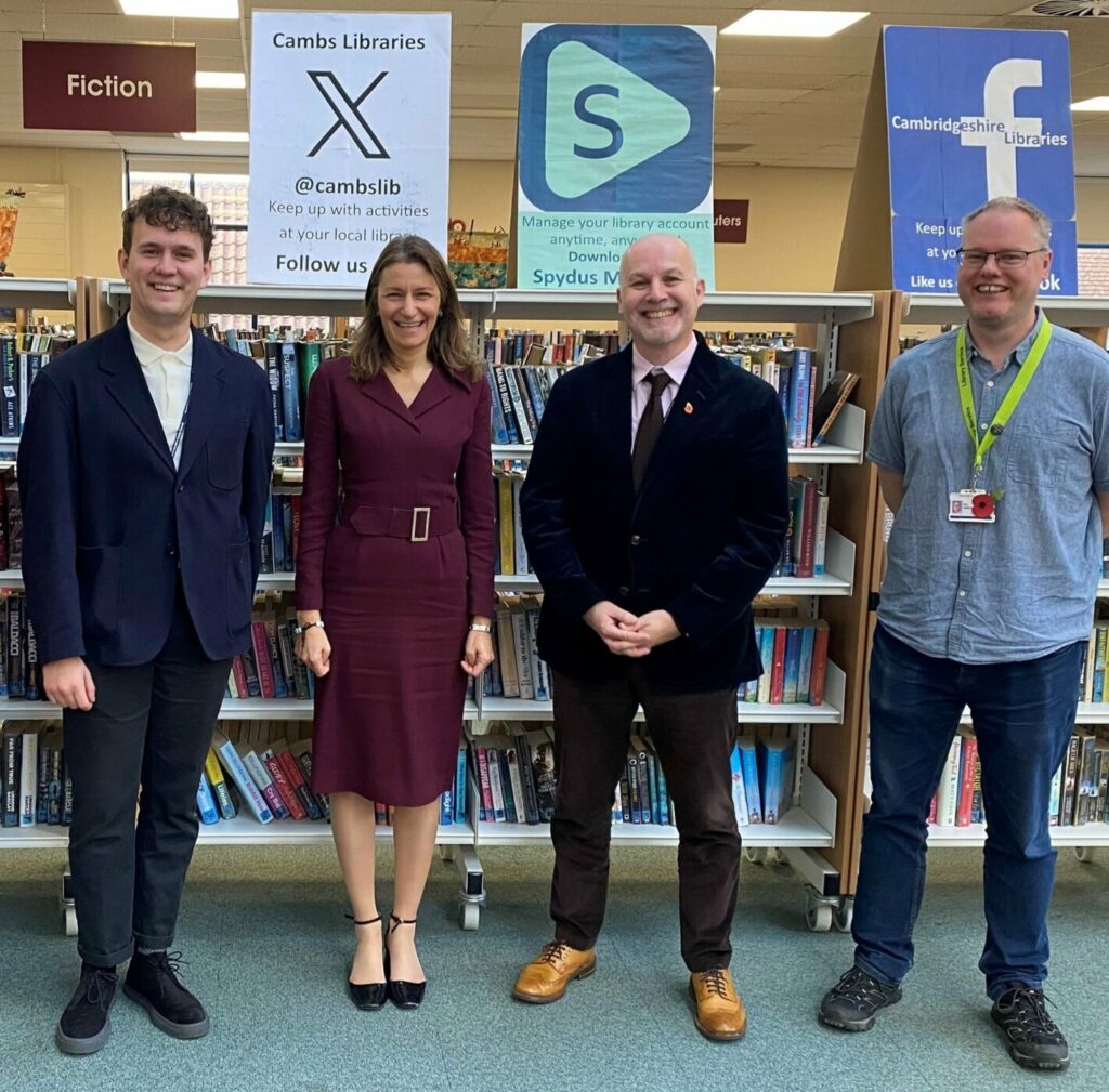 Lucy Frazer MP in Ely Library with representatives from Connecting Cambs, Cambs Libraries & Cambs Skills. Everyone is standing in a row, smiling by bookshelves.