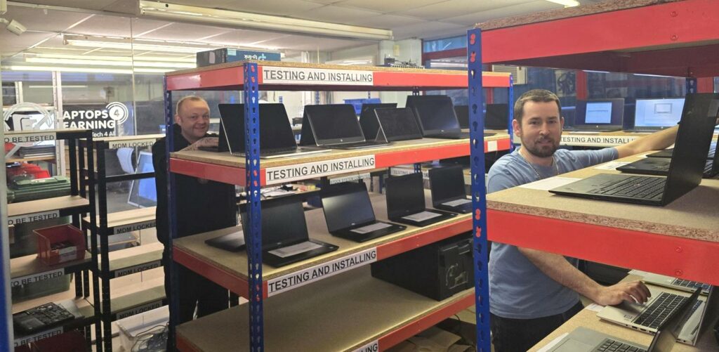 Photo shows shelving units with lots of laptops on, in the Laptops Testing Area in the Sustainable Tech 4 Good offices with two smiling staff members.