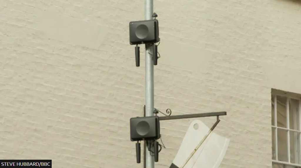 A lamppost with two small cell boxes attached to it, against a brick wall.