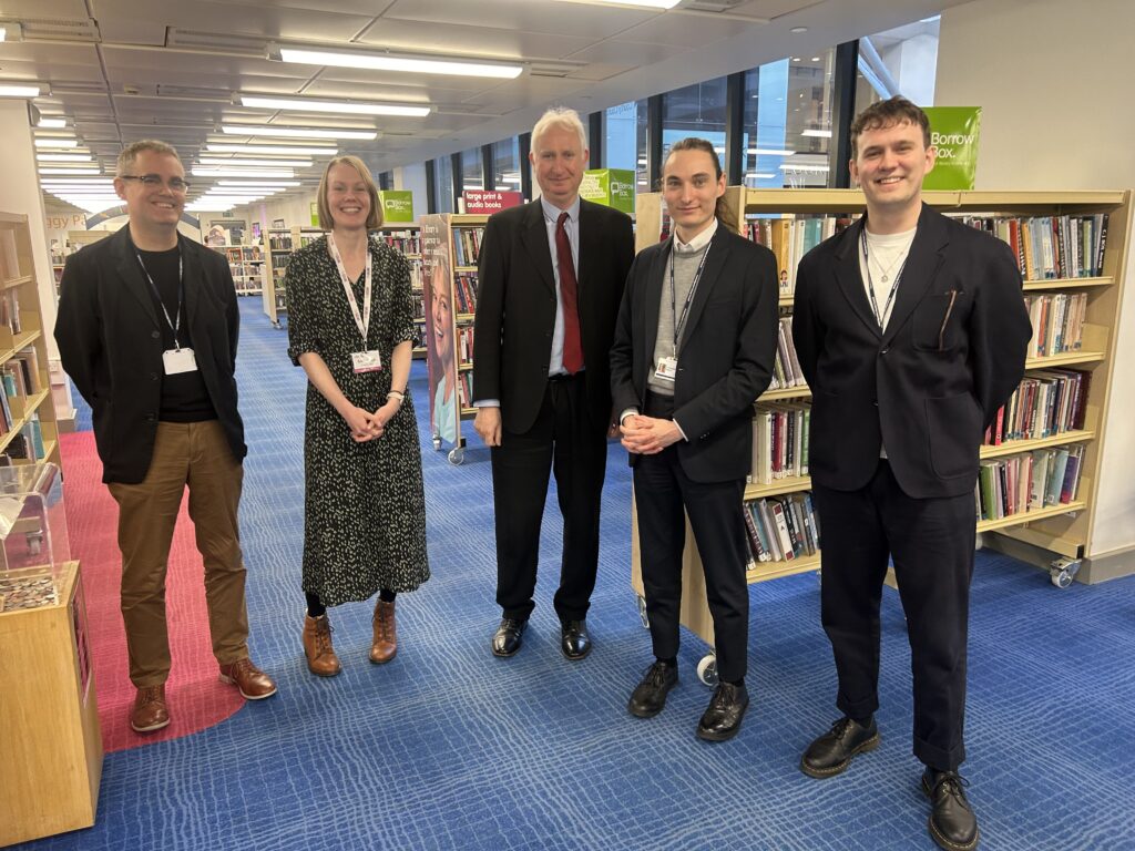 Photo shows Daniel Zeichner MP standing in Cambridge Central Library, in front of some book shelves, with the people he met on the visit. Everyone is smiling and looking happy.