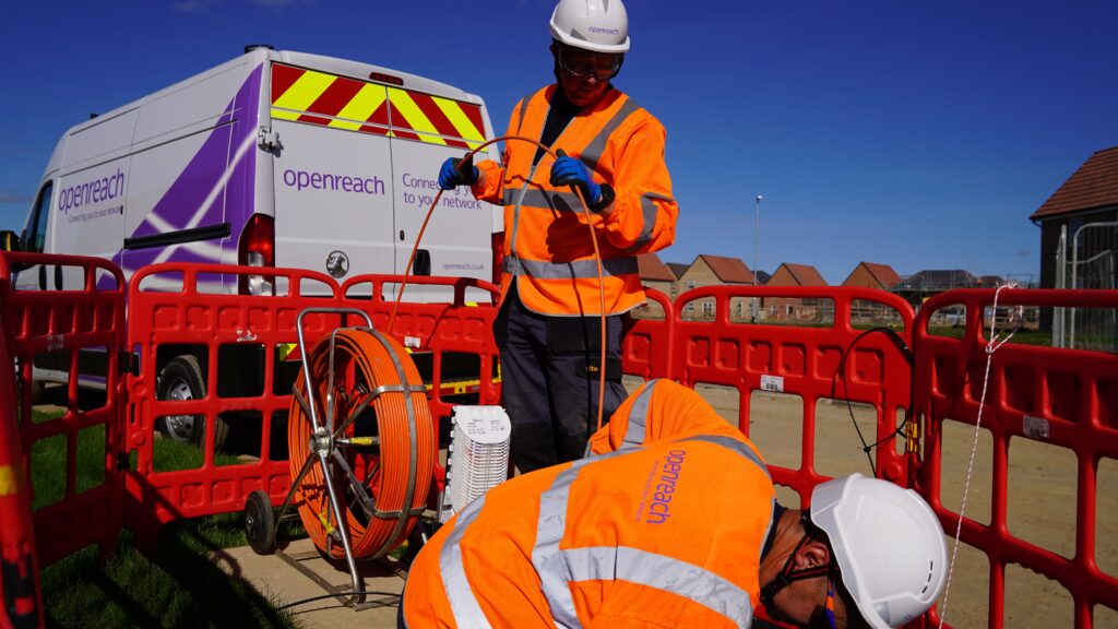 Two Openreach engineers feeding fibre into ducting while standing in front of an Openreach van in a new housing estate on a sunny day.