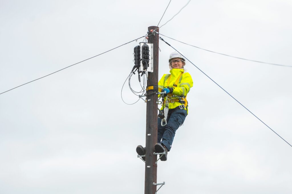 Female Openreach engineer, wearing high-vis and a helmet up a telegraph pole, checking the fibre connections.