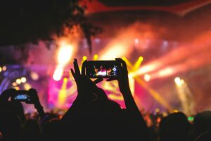 Photo shows a crowded concert venue with someone's hands holding up a smart phone to take a photo of whoever is on stage. There are many other hands in the air in the background also holding phones up to take pictures of the performance.